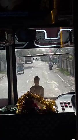 The video shows a view from inside a bus, looking out the windshield. In the foreground, on the dashboard, is a statue of the Buddha adorned with a yellow and gold garland. The bus appears to be on a road in Talpe, Sri Lanka.

The road is asphalt and appears to be a two-lane street with a white dividing line. Several vehicles are visible on the road. To the left, a blue auto-rickshaw is moving away from the viewer. Further down the road, a truck is also moving away. On the right side of the road, a person on a motorcycle is approaching the viewer. Another auto-rickshaw is parked on the right side of the road.

The scene is set outdoors during the daytime. The sky is bright, suggesting it is either morning or afternoon, and the lighting indicates it might be a sunny day. The presence of trees and buildings along the roadside confirms an urban or semi-urban setting.

There are no visible emotions, as the scene is focused on the environment and vehicles. The overall mood is that of everyday travel.

Some text is visible at the top of the windshield, but it is blurry and illegible, except for what appears to be "FM" and "YouTube Channel".