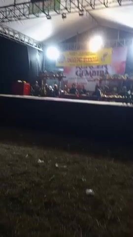 The video shows a nighttime outdoor event with a raised stage covered by a white tent. Bright spotlights illuminate the stage, where a group of people are seated, appearing to be musicians playing traditional instruments. A banner with yellow and red text is displayed behind them. The ground in front of the stage is covered in dirt and scattered with a few pieces of litter. The overall atmosphere suggests a cultural performance or festival.