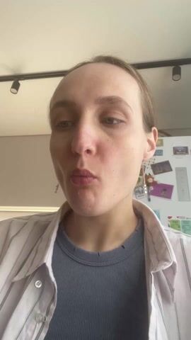 A person eats a spoonful of food, likely in an indoor setting. The person, presumably female, has fair skin, light brown hair pulled back, and is wearing a light pink striped shirt over a blue top. She has long, thin earrings. She looks directly into the camera as she eats. Behind her is a white refrigerator covered with magnets. The lighting is bright, and there are two track lights visible on the ceiling.