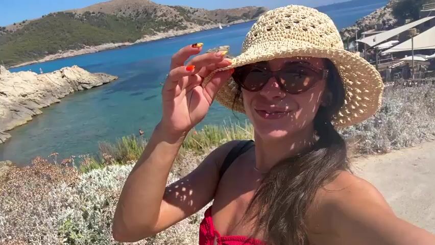 A woman wearing a straw hat and sunglasses is holding the brim of her hat with her right hand. She is smiling and looking at the camera. She is wearing a red bikini top. Behind her, there is a beautiful blue ocean with a rocky coastline and some boats in the water. There are also some people swimming in the ocean. The sun is shining and the weather appears to be warm and sunny. The woman is standing on a dirt path overlooking the ocean.