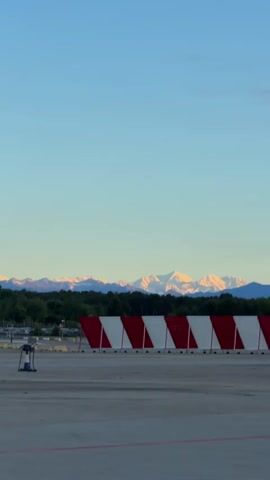 The video shows a view of the Swiss Alps from the Zurich Airport. In the foreground is a grey concrete surface, possibly a runway or tarmac. There's a red and white striped barrier, some trees, a parking lot, and airport infrastructure. The backdrop is a clear, light blue sky with snow-capped mountains in the distance, reflecting sunlight.