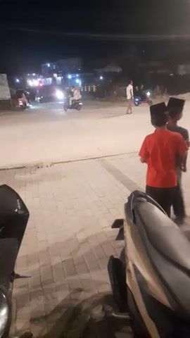 Two young boys wearing caps stand on a paved sidewalk at night, facing a street. One boy is wearing an orange shirt and the other a dark shirt. In the background, vehicles with headlights on are driving on the street. The environment appears to be an urban or semi-urban area with buildings visible in the distance. The foreground shows parts of two motorcycles.