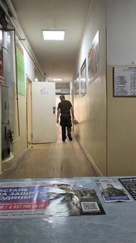 The video shows a long, narrow hallway with beige walls and a drop ceiling with fluorescent lighting. A man wearing a dark uniform, possibly military or security, is walking away from the camera down the hallway. He is carrying a white object in his right hand. The hallway has several doors on the right side and a metal pipe running along the left wall. There are posters on the walls and a bulletin board with a diagram on the far right. In the foreground, there are several printed materials, including what appear to be recruitment posters with images of soldiers and text in Russian. The man is walking towards an open doorway at the end of the hallway. The overall impression is of a utilitarian, institutional environment.
