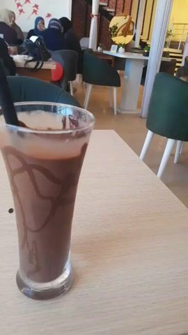 A close-up shot shows a tall glass of chocolate milkshake with chocolate syrup drizzled on the inside walls. The glass is placed on a light-colored wooden table. In the background, out of focus, several people are seated at tables in what appears to be a cafe. The environment is brightly lit with artificial lighting.