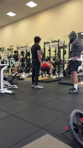 The video captures an indoor gym scene in Los Angeles, United States of America, where several men are engaged in weightlifting. In the foreground, a man in a grey hoodie with "SOHODESIGN LDN" printed on the back and black shorts stands with his hands on his hips, observing the activity, before turning his back to the camera. Another man in a black t-shirt and black pants stands near him. In the midground, a man in a red tank top and dark shorts is performing squats with a heavily loaded barbell within a squat rack, demonstrating intense effort. Further in the background, another man in a light t-shirt and shorts is performing an overhead barbell lift. The gym features rubber flooring, mirrored walls that reflect the interior, and bright overhead lighting. Visible equipment includes multiple squat racks (one with partially visible text "HAMMER STR..."), barbells, and weight plates. A bicycle wheel lies on the floor in the bottom right of the frame, and a couple of water bottles are seen on the floor near the squat rack. The scene depicts a typical training environment with individuals focused on their workout routines.