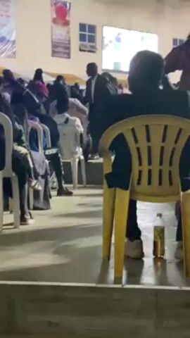 The video shows a gathering of people sitting on chairs in a large, indoor space. They are facing forward, towards a stage area, where a person appears to be speaking. The room seems to be an auditorium or a church, based on the setup and decorations. The people are diverse, with different clothing and hairstyles. Some are wearing masks. The chairs are mostly white and yellow plastic. A water bottle is visible on the floor beside one of the yellow chairs. The environment appears to be well-lit with natural and artificial light. The camera moves slightly, offering different views of the crowd and the overall setting.