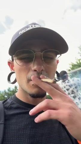The video shows two men outdoors in a park-like setting with trees and foliage in the background. The man in the foreground is wearing a black baseball cap, glasses, and a black t-shirt. He has large ear gauges and piercings on his face. He is looking directly at the camera and appears to be speaking. In the background, another man is performing pull-ups on a set of outdoor exercise equipment. The exercise equipment consists of a metal frame with horizontal bars. To the right of the frame, a bicycle is visible. The ground is covered in gravel and dirt. The lighting suggests it is daytime.