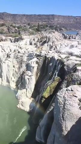 The video shows a wide shot of a large waterfall flowing over a rocky cliff. The water is a greenish color and appears to be turbulent. A faint rainbow is visible in the mist rising from the base of the waterfall. The surrounding environment is rocky and dry, with some sparse vegetation on the hills in the background. The sky is clear and blue. The camera is positioned above and to the side of the waterfall, providing a dramatic perspective.