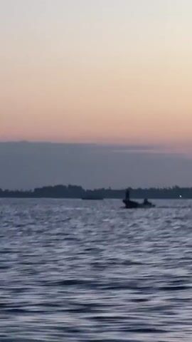 A video captures a wide shot of a body of water, likely the ocean, under a sky transitioning from dusk to night. The water exhibits moderate waves. A small, dark boat with what appears to be two individuals aboard is visible in the middle ground, moving from left to right across the frame. The boat is small and dark, and the figures on it are indistinct silhouettes. The background features a faint, dark landmass, possibly an island or coastline, silhouetted against the horizon. The sky displays soft hues of pink and orange near the horizon, fading into a muted gray higher up, with scattered, wispy clouds. The overall atmosphere is serene and calm, suggesting an early morning or late evening setting.