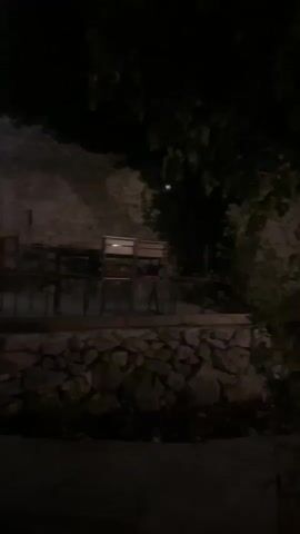 The video shows a nighttime scene outside a building with stone walls. There are tables and chairs set up on a patio area. An arched doorway leads into the building, and stairs are visible inside. Green foliage hangs from the building's exterior, and red lights are visible through windows on the upper level, casting an eerie glow. The area is dimly lit, with spotlights illuminating sections of the stone wall.