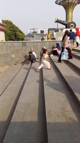 The video shows an outdoor scene with a group of people walking up a set of wide concrete stairs. In the background, there is a large, golden, trumpet-shaped structure with a cannon mounted in front of it. A palm tree is also visible. The people in the foreground are dressed casually, and some are wearing hijabs. A woman is carrying a white plastic bag. Children are also present. The stairs are well-lit by sunlight. Some colorful toys are displayed on the side of the stairs at the bottom of the frame.