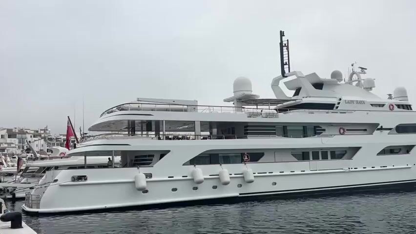The video shows a large white yacht docked in a marina. The yacht is named "LADY HAIA" and has multiple decks and windows. There are several buoys attached to the side of the yacht. In the background, other boats and buildings are visible. The sky is overcast.