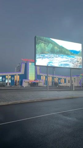 A wide shot captures a modern building with blue and white accents, illuminated by blue and yellow lights. A large digital billboard in front of the building displays a scenic image of a river flowing through a mountainous landscape. The sky is overcast and dark. The foreground shows a wet asphalt road and a paved walkway. The building appears to be a train station or a similar public facility. The time stamp indicates the video begins at 00:00 and ends at 00:04. A person is briefly visible walking towards the building.