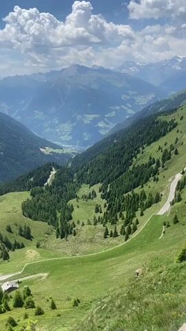 A sweeping, vertical drone shot captures a majestic mountain landscape on a clear day. The foreground is dominated by a steep, grassy slope with scattered trees. A winding paved road cuts through the middle ground, revealing a few small buildings and solar panels. The road leads into a lush green valley dotted with dense evergreen trees. In the distance, a series of imposing, jagged mountain ranges recede into the hazy blue atmosphere. White, puffy clouds drift across the bright blue sky, casting shadows on the mountain peaks. The scene is one of serene natural beauty and vastness.