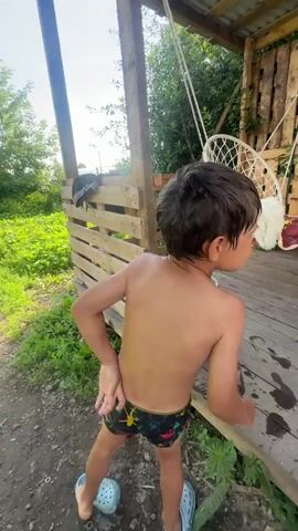 This short video clip captures a young boy, likely pre-school age, enjoying himself outdoors on a sunny day. He is shirtless and wearing swim trunks and blue clogs. The boy is in the process of playing with water, evidenced by his wet hair and the drops of water visible on his skin. In the background, a blue bucket, possibly containing water, sits on a wooden porch. The setting appears to be a rural or semi-rural area, with greenery and wooden structures suggesting a backyard or a dacha. A wooden shed and a hanging chair are also visible on the porch, hinting at a leisure or recreational space. The boy's expression is joyful and animated, with a wide smile and playful movements, indicating he is having a good time. The overall atmosphere is one of carefree childhood play on a warm day. The sky is clear and blue, suggesting fair weather.
