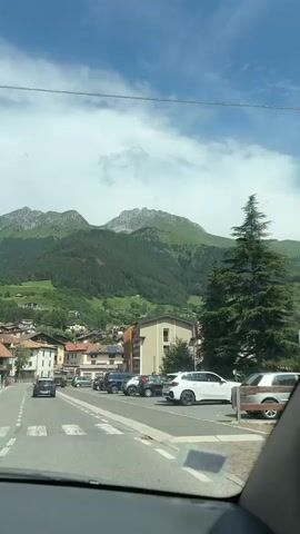 The video shows a view from inside a vehicle driving on a road in a town at the base of a mountain. The road has painted lane dividers and a pedestrian crossing. Several cars are parked on the side of the road and others are driving on the road. The town consists of multi-story buildings with tiled roofs. The mountain is covered in trees and some rocky peaks are visible at the top. The sky is blue with scattered white clouds. The lighting suggests it is daytime, likely late morning or early afternoon.