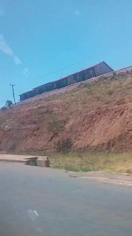 The video shows a view from a moving vehicle. A silver van with a red and yellow design drives past. The camera then focuses on a hillside with a building and a water tower on top. The sky is clear and blue. A road is visible in the foreground. The video seems to be shot during daytime in a sunny location.