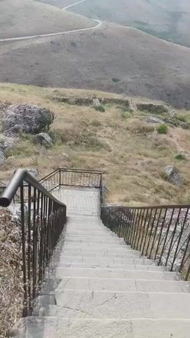 A stone staircase with metal railings descends a grassy hillside dotted with rocks and the ruins of stone structures. Beyond the hillside, a winding road snakes through rolling, barren hills under a hazy sky. The camera pans slowly to the right, revealing a vast, mountainous landscape.