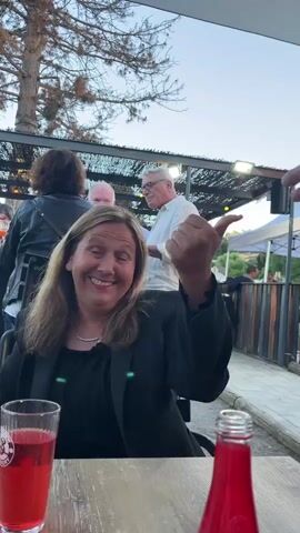 The video captures a social gathering at an outdoor venue, possibly a restaurant or bar, with a setting sun casting a warm glow. In the foreground, a woman with blonde, shoulder-length hair, wearing a black blazer over a dark top, is seated at a wooden table. A glass of red beverage with a white logo is in front of her. She is smiling and looking upwards. To her right, another glass and a bottle with a red liquid are visible on the table.

In the background, several other people are present. To the left, a woman with dark hair and a black top with white polka dots is seated, also smiling and looking towards the camera. Further back, a woman with dark curly hair and a black leather jacket is standing. A man with white hair and a white shirt is seen clapping. Another man with glasses and a white shirt is also visible. The environment appears to be an open-air structure with a roof and some lighting fixtures. Trees and foliage are present in the upper portion of the frame. The overall atmosphere suggests a relaxed and convivial occasion.