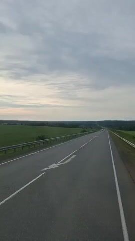 The media file captures a view from a vehicle traveling along an asphalt road. The road stretches into the distance, with white lines marking the lanes and a metal guardrail bordering the left side. Beyond the guardrail, expansive fields of green crops stretch out, with a line of trees forming the horizon. The sky is overcast with a mix of grey and white clouds, suggesting a potentially cool or damp day. Sunlight filters through the clouds, creating a soft, diffused light.

Several cars are visible on the road, indicating a moderate level of traffic. The vehicles are moving in the same direction as the viewer. The overall setting is rural, characterized by open fields and a clear, unobstructed view of the landscape. The time of day appears to be either early morning or late afternoon, given the soft lighting and the relatively low angle of the sun, though the overcast conditions make it difficult to be precise.

The mood evoked is one of travel and journey, with the open road and the vast landscape suggesting movement and exploration. There are no discernible people in focus, but the presence of cars implies human activity. The location is identified as Kuregovo, Russia, which aligns with the broad, agricultural landscape depicted. There is no visible text within the frame.