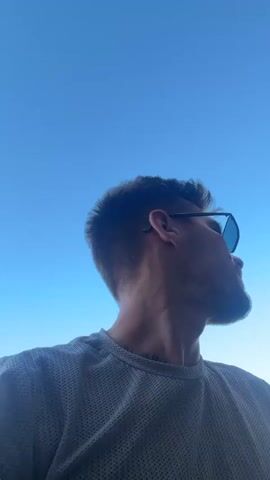A man with short brown hair and a beard is shown from the chest up. He is wearing sunglasses and a grey textured t-shirt. The video appears to be a selfie taken outdoors with a bright blue sky in the background. In some frames, a building with a pink and cream colored facade and a blue-tinted window is visible. There is also a roofed structure with a clear canopy overhead.