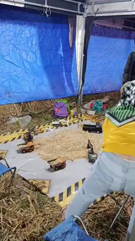 The video shows an outdoor scene at night. In the foreground, a person wearing a yellow jacket and light blue jeans is crouched down, facing away from the camera. Their back is to a large pile of what appears to be grain or seeds. Several toy dump trucks are arranged around the pile on a gray tarp with yellow caution stripes. A purple bag is visible behind the pile. To the right of the foreground person, another person wearing a brown hijab and light-colored pants is standing and looking to the left. They are holding a phone in their right hand. Behind them, there is a tent-like structure with blue tarps on the left and white and black material on the right. In the background, a colorful carousel-like amusement ride is lit up. Several other people are visible in the background, some standing near the ride. The ground is covered in dry grass and straw.