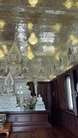 The video shows an interior room in a temple with a large silver-colored Buddha statue as the focal point. The statue sits on a multi-tiered platform adorned with floral arrangements and smaller statues. Two standing monk statues flank the Buddha, and two seated monk statues are positioned in the foreground. The room features dark wood paneling and a polished wood floor. The ceiling is covered with reflective silver-colored tiles, creating a shimmering effect that mirrors the light from several crystal chandeliers. A small table with a lamp sits on the left side of the frame.