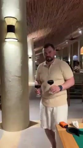A man is standing in an upscale restaurant, holding two glasses of red wine. He has short brown hair and a beard, and is wearing a tan polo shirt, white shorts, and a blue smartwatch. The restaurant has a thatched ceiling and white pillars, and is dimly lit by lamps. He raises the glasses in a toast, then turns and walks away, carefully carrying the wine. The camera follows him, showing other tables and patrons in the background. He turns back towards the camera for a moment, before continuing to walk.