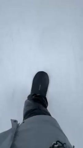 The video shows a first-person perspective of someone snowboarding on a snow-covered slope. The snowboarder is wearing dark pants and boots, and the snowboard itself is black. The snow is mostly white with visible tracks. In the background, there are other snowboarders and skiers, as well as a building that looks like a ski lodge. The weather seems cold and the wind is audible in the recording. The snowboarder is moving downhill.