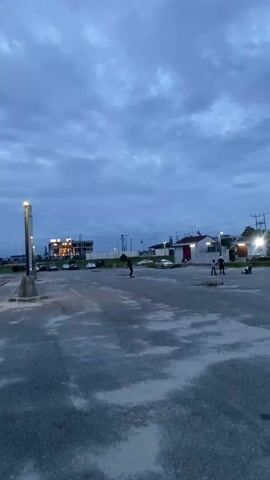 In a wide, stationary shot, a young person skateboards across an empty parking lot. It is dusk, with a clouded sky and street lights. The skater wears black pants, a dark chest harness, and gloves. In the background are parked cars, a low building, and utility poles. Other people are visible standing in the distance.