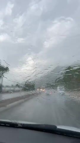 The video shows a view through a wet car windshield as a car drives on a road in the rain. Raindrops streak down the windshield, and the wipers are visible in the foreground. Other cars can be seen on the road ahead, with their headlights and taillights blurred by the rain and the windshield's condition. The sky is overcast and gray, indicating heavy rain. The environment appears to be a highway or a similar type of road, with trees and possibly buildings visible in the background. The overall impression is of driving in adverse weather conditions.