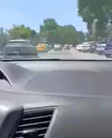 A first-person perspective video captures a view from inside a vehicle driving on a street. The road appears to be a multi-lane urban street lined with trees and buildings. Several vehicles are visible, including yellow taxis, sedans, and a motorcycle. The camera is focused on the road ahead, showing traffic and an intersection. The lighting suggests it is daytime with a clear sky. The video is short and appears to be a snippet of a longer recording, with no apparent events of note occurring within this timeframe.