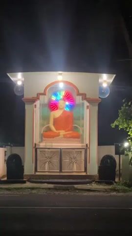 A statue of Buddha sits within an alcove on a wall at night. The Buddha statue is orange and depicted in a meditative pose. A circular halo of colorful lights pulses behind the statue's head, cycling through red, orange, yellow, green, blue, and purple. The alcove is framed by architectural elements, with two light fixtures above and two closed metal doors below the statue. Flanking the alcove on either side are dark, indistinct objects, possibly statues or decorative elements. The overall scene is lit by the statue's halo and the overhead lights, casting some shadows and illuminating the immediate surroundings. The area appears to be outdoors, possibly near a road or walkway, with some vegetation visible to the right of the alcove. The atmosphere is quiet and contemplative, highlighted by the nighttime setting and the illuminated Buddha.