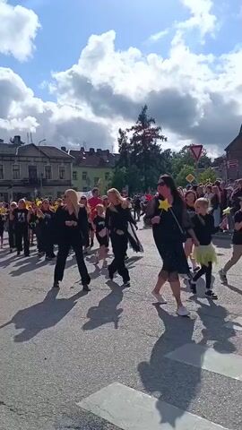 A group of people, many in black clothing, are walking down a paved street. Some are carrying yellow star-shaped objects and balloons. The sky is blue with fluffy white clouds. Buildings line the street in the background, and trees are visible. The sun is casting long shadows.