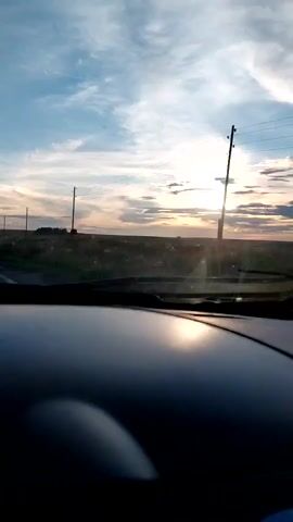 The video shows a view from inside a vehicle looking out the windshield at a sunset. The sky is partly cloudy with the sun visible low on the horizon. The landscape appears to be flat and rural, with fields and utility poles. The dashboard of the vehicle is visible in the foreground, reflecting the light from the sunset. The video is grainy and appears to be of low resolution.