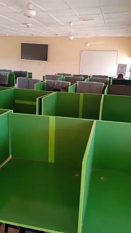 The video shows an empty computer lab. The lab is filled with rows of individual computer workstations, each separated by green partitions. There are monitors at each station, mostly covered by a dark grey material. The room is brightly lit with several ceiling fans and a drop ceiling with rectangular panels. A large flat screen television is mounted on one wall, and a whiteboard is on another. One person is sitting at a computer in the back of the room, but the rest of the lab is empty.