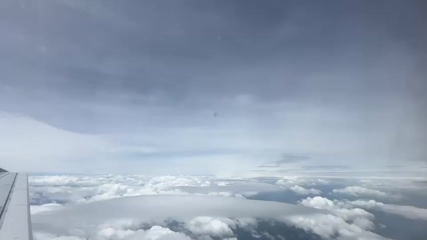 This is a view from an airplane window overlooking a vast expanse of clouds. The airplane wing is visible in the lower left corner. The sky above is a mix of blue and grey clouds, with streaks of lighter clouds suggesting wind movement. Below, a dense layer of white clouds stretches to the horizon, with some of the clouds appearing flattened and layered, possibly lenticular clouds. The light suggests it is daytime.