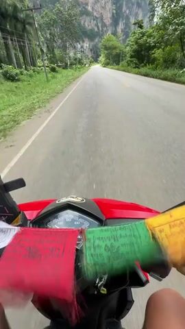 The video shows a first-person perspective of someone riding a red scooter or motorcycle down a paved road. The rider’s hands are visible, and prayer flags in red, green, and yellow are tied to the handlebars. The road is flanked by lush green grass and trees on the left, and towering, rugged cliffs covered in vegetation on the right. Power lines are visible along the side of the road. The sky is partly cloudy. The road stretches ahead into the distance, with another motorcycle visible in the far distance. The overall impression is of a scenic, outdoor journey through a natural landscape.
