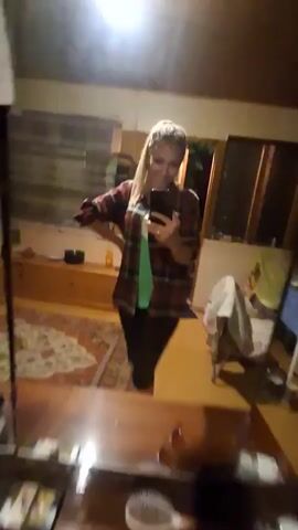 In this media file, a woman is taking a selfie in a room, likely a bedroom or living area, reflected in a mirror. She is wearing a red and black plaid shirt unbuttoned over a green t-shirt and black pants. Her blonde hair is pulled back in a ponytail, and she is smiling at the camera while holding her phone. She has a ring on her left hand.

The setting appears to be indoors, with wooden furniture visible in the background, including a dresser or cabinet. A rug with a patterned design is on the floor. There is a window with blinds in the background, and a bright light source, possibly a ceiling light, is illuminated. The overall lighting suggests it might be evening or nighttime, as the interior light is prominent.

The woman is engaged in the activity of taking a photograph of herself. Her posture and smile convey a sense of casualness and happiness. There is no visible text in the media file. The location context provided is Dachnaya, Russia.