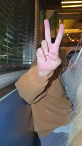 The video shows a person with long blonde hair partially blocking the view of a person making a peace sign. The person making the peace sign is sitting on a blue bench inside a building near a window with blinds. The peace sign is being made with the right hand, and the person making the sign is wearing a brown sweatshirt. Another person is sitting next to them. There is an illuminated area visible outside the window.