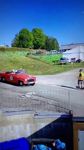 A red vintage convertible with a blue number 20 sticker on the door drives by on a paved road. The car has two people in it, a driver and a passenger. A golf cart is parked on the right side of the road. A person wearing a yellow safety vest stands on the right side of the road. In the background, there are trees, a white tent, and several other cars parked on a grassy area. The sky is blue with a few clouds. The scene appears to be a sunny day event.