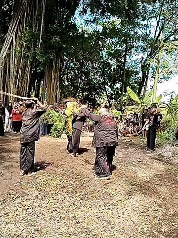 A group of people is gathered in an outdoor area, with trees visible in the background. In the center of the scene, several individuals dressed in black martial arts attire are engaged in a sparring match. One person is thrown to the ground during the altercation. There is a crowd of spectators watching the events unfold. The ground is covered with fallen leaves.