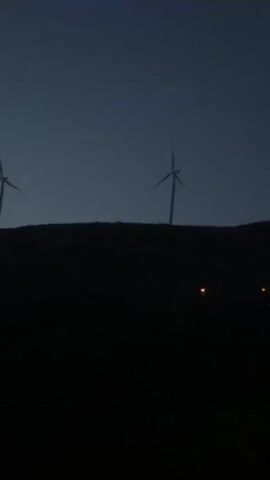 The video shows two wind turbines silhouetted against a dark blue sky. The turbines are on a hilltop, which is also in silhouette. There are a few faint lights visible below the hilltop. The turbines' blades turn intermittently. The shot is static.