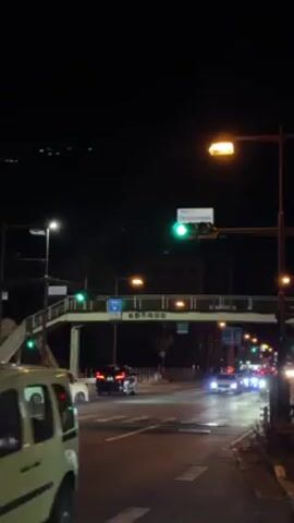 The video shows a street scene at night. Cars are driving on the road, and traffic lights are visible, including a green light. A pedestrian overpass spans the road with signs. The sky is dark. Streetlights illuminate the area.