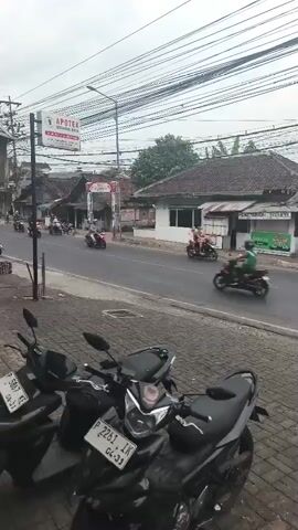 The video shows a street scene from the perspective of someone standing on a sidewalk. Several motorcycles are visible, some parked on the sidewalk in the foreground and others riding on the street. There is also a light-colored van and a red car driving on the road. Buildings line the street, and there are many overhead wires. In the background, there are trees and hills. A large red and white banner hangs over the street. People are seen riding on the motorcycles, and some are walking. The sky is overcast.