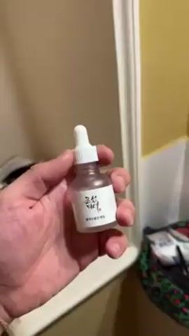 A person places a small bottle with a dropper on a shelf. The person then takes the bottle and, looking into a mirror, applies the liquid to their face. The setting is a bathroom.