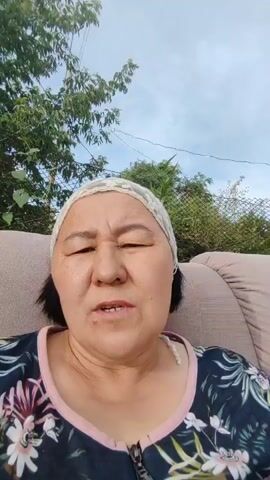 A woman wearing a headscarf and a floral patterned shirt is speaking directly to the camera. She appears to be sitting outdoors with trees and a cloudy sky visible in the background. She is looking up and to the left with a serious expression and her mouth is slightly open as if she is in the middle of speaking.