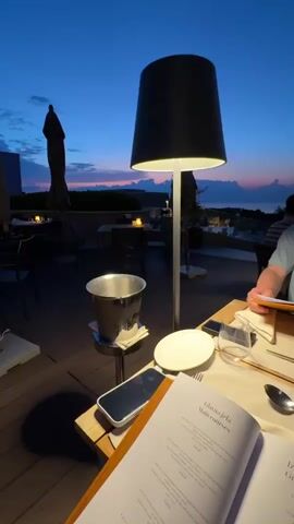 The media file shows a close-up view of a table at an outdoor restaurant at dusk. The table is set for a meal with a menu open, a glass of water, silverware, and a white plate. An ice bucket sits on a small side table nearby. A large, modern lamp with a black shade illuminates the scene. In the background, the sky is a deep blue with hints of pink and orange, suggesting a sunset. Silhouettes of umbrellas and restaurant furniture are visible, along with a person sitting at another table to the right. The overall atmosphere is calm and sophisticated, possibly indicating a relaxed evening dining experience.

The text visible on the menu includes "Glavna jela Main courses" and "Izbor s roštilja Grill selection". The visible text is in English and Croatian. The location is implied to be Crveni Vrh, Croatia, suggesting a coastal setting.
