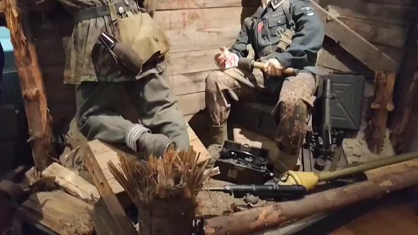 The video shows a museum exhibit depicting a World War II scene. Two mannequins dressed in military uniforms are positioned in a makeshift bunker made of wood and sandbags. The mannequin on the left is standing and facing away from the camera, wearing a camouflage uniform and carrying a satchel and what appears to be a canteen. The mannequin on the right is seated, facing the camera, wearing a grey uniform with insignia, a helmet, and patterned camouflage trousers. This mannequin is holding a grenade in its right hand and appears to be in a relaxed or contemplative pose. Various military equipment is displayed around the mannequins, including a radio, a machine gun, a fuel can, and a shovel or tool. The environment is dimly lit, emphasizing the rough and weathered look of the bunker.