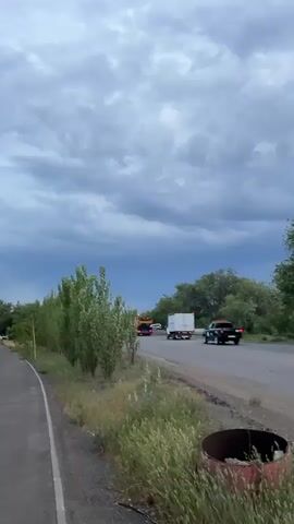 A time lapse video shows a road with several vehicles driving on it, including a truck and a car. The sky is cloudy and appears to be darkening, suggesting an approaching storm. Trees and vegetation line the side of the road.